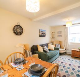 A living room with a wooden dining table and chairs a green sofa beige armchairs a clock on the wall and a window with curtains at Guinea Cottage in Brewers Quay Harbour