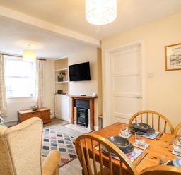 A living room and dining area with a wooden dining table set with bowls and glasses a fireplace mounted tv armchairs a window with curtains and a rug on the floor at Guinea Cottage Brewers Quay Harbour
