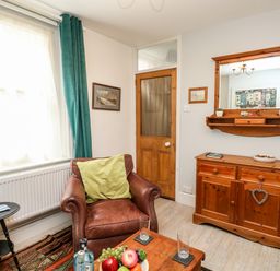 A living room with a brown leather armchair and green cushion a wooden cabinet with a mirror above and a wooden table with fruit and bottles at Harbour Catch Cottage in Weymouth