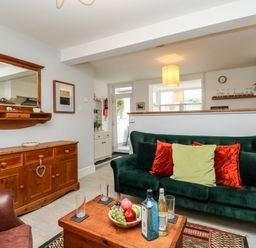 A living room with a green sofa with red and green cushions a wooden coffee table with bottles and a fruit bowl and a wooden sideboard with a mirror at Harbour Catch Cottage in Weymouth