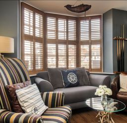 A sitting area with a striped armchair, gray sofa, round glass coffee table, bedside lamp, and wooden window shutters at Harbourside House in Brewers Quay Harbour