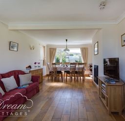 A living room with a red sofa cushions a dining table with wooden chairs and a television on a wooden stand at Hillview Bungalow in Burton Bradstock