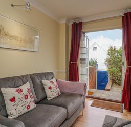 A living room with a gray sofa and red floral cushions next to an open door with red curtains at Hops House in Brewers Quay Harbour