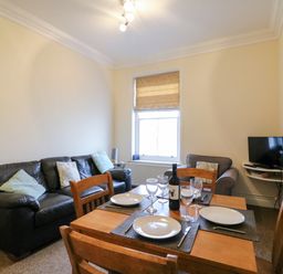 A dining table with plates and wine glasses beside a black sofa and an armchair with a television on a wall shelf at Harbour Watch Apartment 6 Brewers Quay Harbour