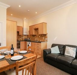 A kitchen and dining area with wooden cabinets and a table set with plates and glasses next to a black sofa at Harbour Watch Apartment 6 in Brewers Quay Harbour