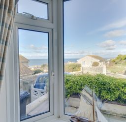 A window with plaid curtains overlooking a garden hedge a model sailboat and houses with a sea view at Langham Cottage in Portland