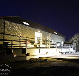A stone cottage with a wooden railing and exterior lights at night at Leys At Valley View Farm in Uplyme