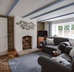 A living room with a stone fireplace, sofas with cushions, a TV on a wooden cabinet and patterned curtains at Lilac Cottage in Burton Bradstock