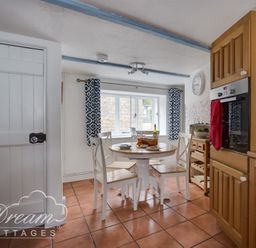 A kitchen with a round table and four chairs near a window with blue patterned curtains at Lilac Cottage Burton Bradstock