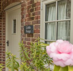 An exterior view of a brick house with a cream door and window a black sign reading Little Forge and a pink flower in foreground at Little Forge in West Lulworth