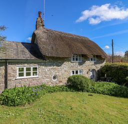 A stone cottage with a thatched roof and garden with bushes and flowers at Lychgate Cottage in Osmington