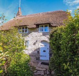 A stone cottage with a thatched roof and a white arched door surrounded by green bushes at Lychgate Cottage in Osmington