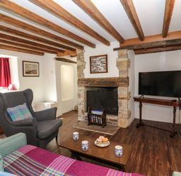 A living room with wooden beams a fireplace an armchair a sofa a wooden coffee table with mugs and cake and a television at Lychgate Cottage in Osmington
