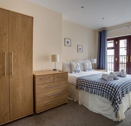 A bedroom with a wooden wardrobe and chest of drawers beside a bed with pillows and folded towels near a window with curtains at New Boathouse in Weymouth