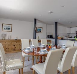 A dining area with a wooden table set for eight and white chairs next to a kitchen with white cabinets and black countertops at Old Coastguard Apartment 2 Weymouth