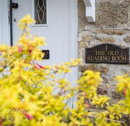 A white door with a decorative window and a plaque reading The Old Reading Room on a stone wall with yellow flowering plants in front at The Old Reading Room in Langton Herring