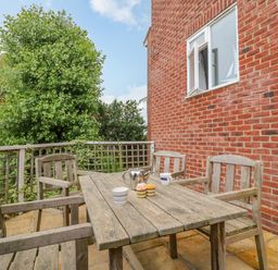 An outdoor patio with a wooden table and four wooden chairs a teapot and cups on the table next to a red brick house with a window at Ocean Watch 1 Greenhill