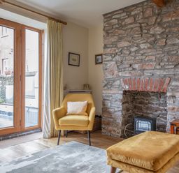 A sitting area with a yellow armchair and matching footstool next to a stone fireplace and wooden framed glass doors at 14a Courtenay Street Salcombe