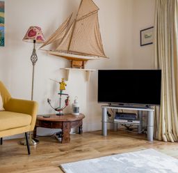 A living room with a yellow armchair a floor lamp a model sailboat on a wall shelf a wooden coffee table a television on a stand and curtains at 14a Courtenay Street Salcombe