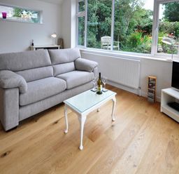 A living room with a grey sofa a glass top table with wine and glasses a television and wooden floor at Pebbles Burton Bradstock