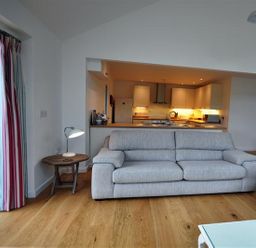 A living room with a gray sofa a wooden side table with a lamp and an open kitchen in the background at Pebbles in Burton Bradstock