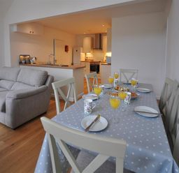 A dining table set with plates mugs and glasses of orange juice next to a living area with sofas and an open kitchen in Pebbles Burton Bradstock