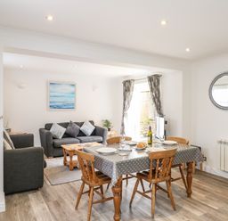 A dining area with a table and wooden chairs adjacent to a living room with sofas and a painting at The Pines in Weymouth