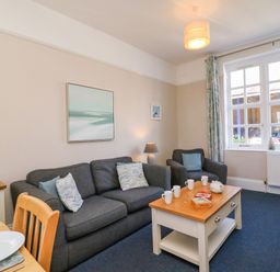 A living room with a dark sofa and armchair with cushions a wooden coffee table with mugs and games a window with curtains and a lamp at Reaver in Brewers Quay harbour