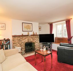 A living room with a beige sofa black armchair stone fireplace television and red carpet at River Cottage in Burton Bradstock