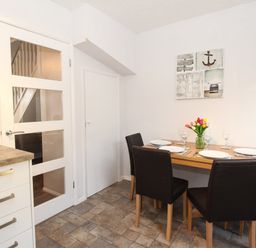 A dining area with a wooden table set for four people with plates glasses and a vase of flowers with a door and kitchen counter nearby at Rockpools Weymouth