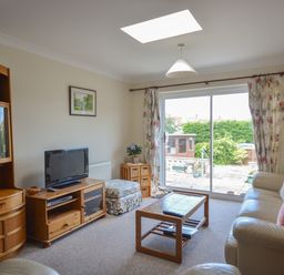 A living room with a sofa, coffee table, television on a wooden stand, floral curtains, and sliding glass doors opening to a garden at Sanjernin in Swanage