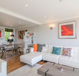 A living room with a white sectional sofa with colorful cushions a wooden coffee table a dining table with chairs and wall art at Sea King House in Cove Castle