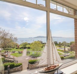 A window view of a park with people sitting and walking near the sea with boats sailing and a model sailboat on the windowsill at Sea King House in Cove Castle