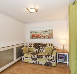 A small room with a floral patterned sofa a side table with a lamp and board games a wall painting and a green curtain at Sea Pebbles in Weymouth