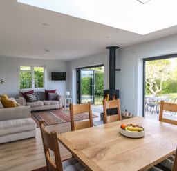 A dining table with chairs and a bowl of fruit in an open living room with sofas a wood stove and doors opening to a garden at Shipton Hill View in Shipton Gorge