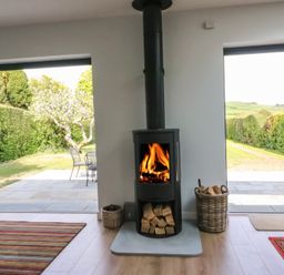 A wood burning stove with fire inside and logs stored underneath and next to it in a room with open doors showing a garden at Shipton Hill View in Shipton Gorge