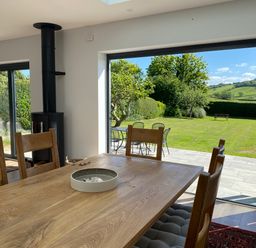 A dining area with a wooden table and chairs next to large open doors leading to a garden with patio furniture and greenery at Shipton Hill View in Shipton Gorge