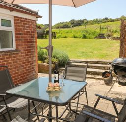 An outdoor patio with a glass table and chairs a bottle of wine and cupcakes on the table a barbecue grill and a grassy garden area at Studland Cottage in Studland