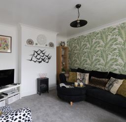 A living room with a black sectional sofa with patterned cushions a wooden shelving unit and a tv on a white stand at Sunny Days in Weymouth