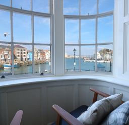 A bay window with armchairs and cushions overlooking a harbor with boats and buildings at Tradewinds Brewers Quay Harbour