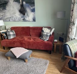 A red sofa with cushions and a gray footstool with a folded blanket in a living room with wooden floor and a large photograph of a lighthouse at Tradewinds Brewers Quay Harbour