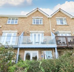 A rear view of a brick house with a glass balcony and patio doors surrounded by bushes at Waters Edge in Wyke Regis
