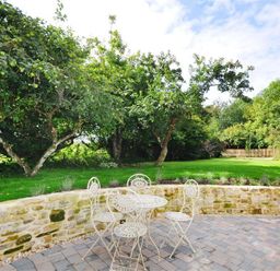 A garden patio with a stone wall and metal table and chairs set surrounded by trees and grass at Warmwell Lodge in Warmwell
