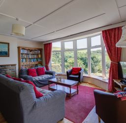 A living room with gray sofas and red cushions a wooden coffee table a bookshelf a large window with red curtains and a floor lamp at Waterson in Osmington