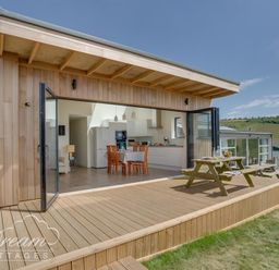 A wooden deck with picnic tables outside a cottage with open glass doors showing a kitchen and dining area at The Wave in Eype