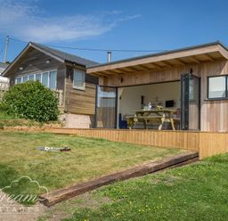 A wooden cottage with open glass doors leading to a dining area and a grassy yard with toys at The Wave in Eype