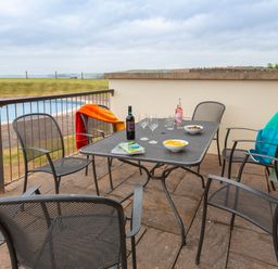 An outdoor patio with a metal table and six chairs with drinks bowls and blankets near a body of water at 2 Ocean's Edge in Thurlestone
