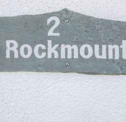 A rectangular slate house sign with the text 2 Rockmount mounted on a white textured wall at 2 Rockmount Salcombe