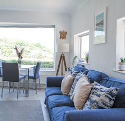 A sitting room with a blue sofa and dining table with grey chairs near large windows at 2 Rockmount in Salcombe