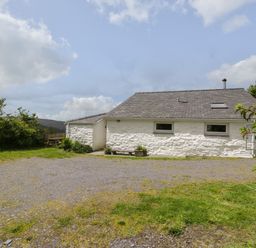 A white stone cottage with a gray roof and small windows surrounded by grass and bushes at Y Bwthyn Elidir Fach Mountain near Llanberis